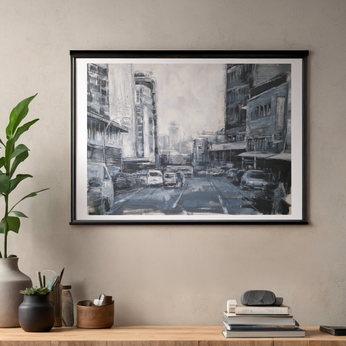 A framed black and white city street art print hangs on a textured beige wall above a wooden shelf with books, a plant, and small decorative vases.