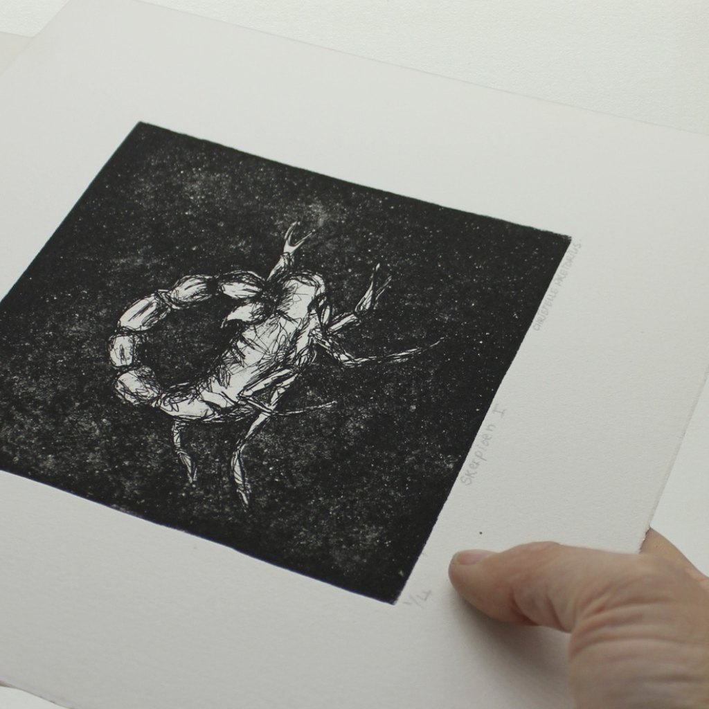 A black and white ink print of a scorpion on a white sheet of paper, held by a hand at the bottom right corner.